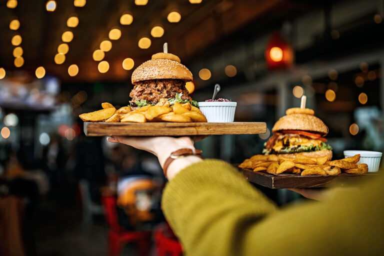 Two burgers with fries being carried on a platter in a restaurant with bokeh lights in the background