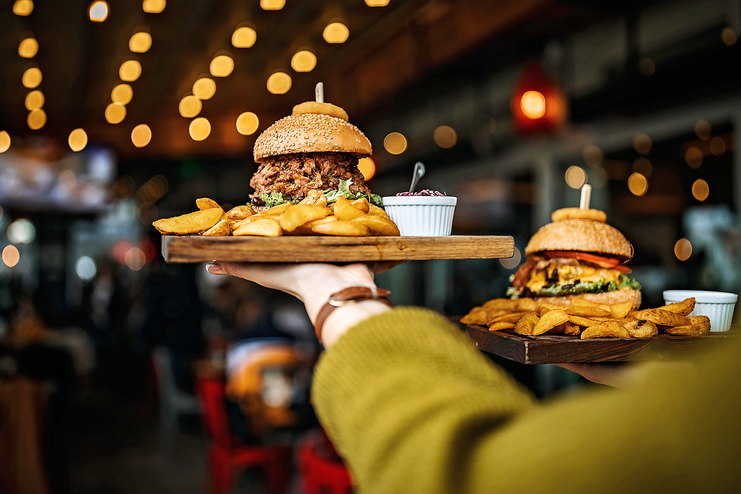 Two burgers with fries being carried on a platter in a restaurant with bokeh lights in the background